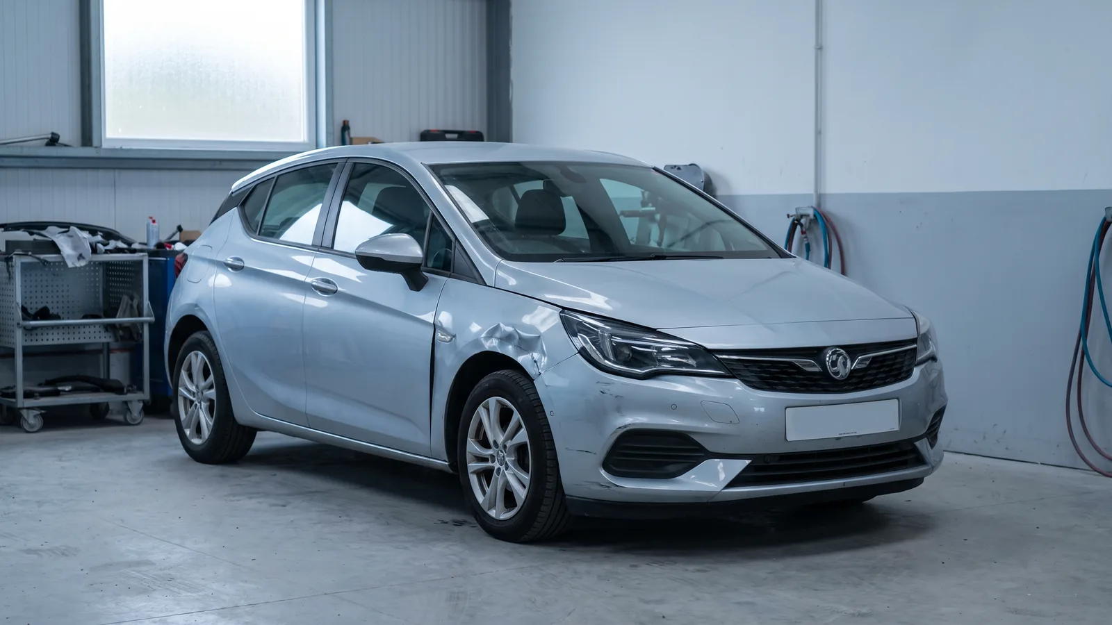 A silver hatchback with damage to its front wing, inside a vehicle body repair workshop